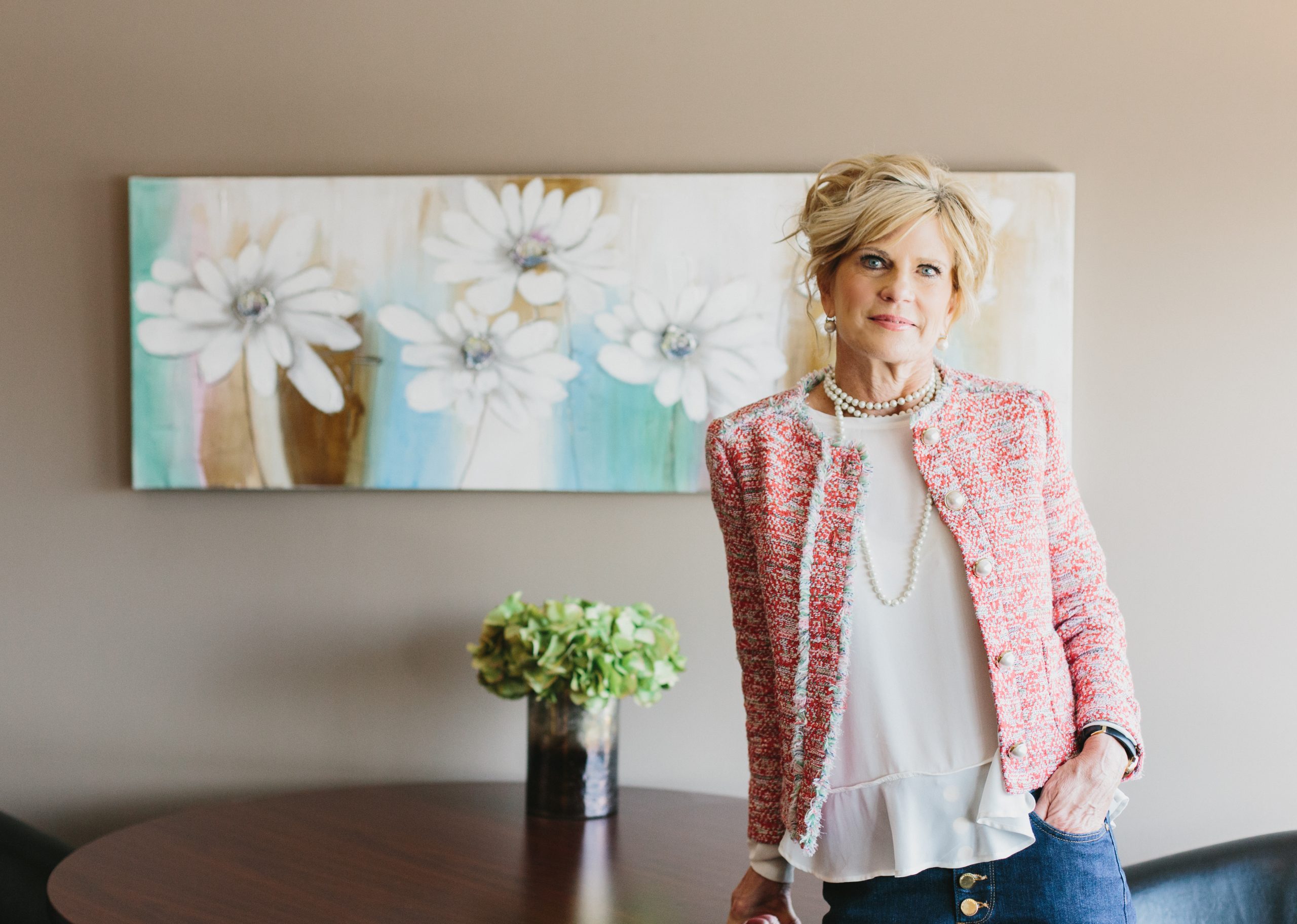 Mary Jane James stands, leaning on a desk. She's wearing a pink blazer over a white top.