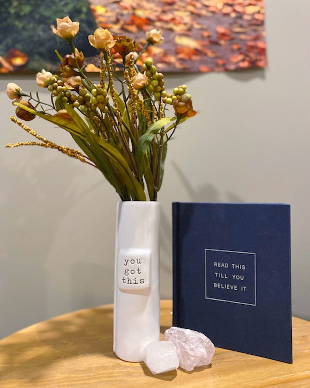 A table with a white vase with "you got this" printed on it with flowers, two crystals, and a navy blue book with "read this till you believe it" printed on the cover.