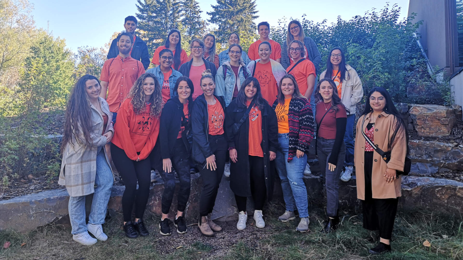 A group of smiling SACE staff in orange shirts stands together in front of a monument at Fort Edmonton Park.
