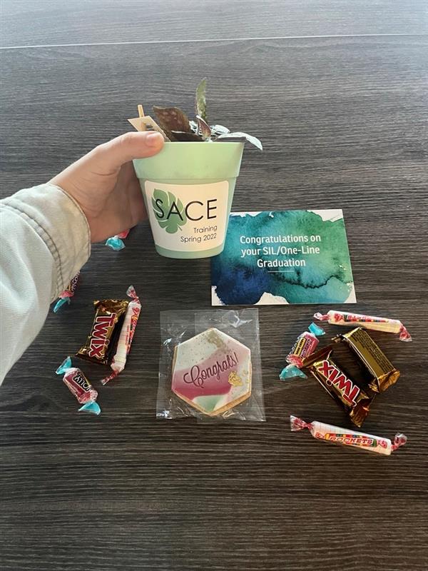 Graduate of the volunteer services program SIL training holds up assorted gifts: a hand-decorated flower pot, candies, cookies, and a postcard.