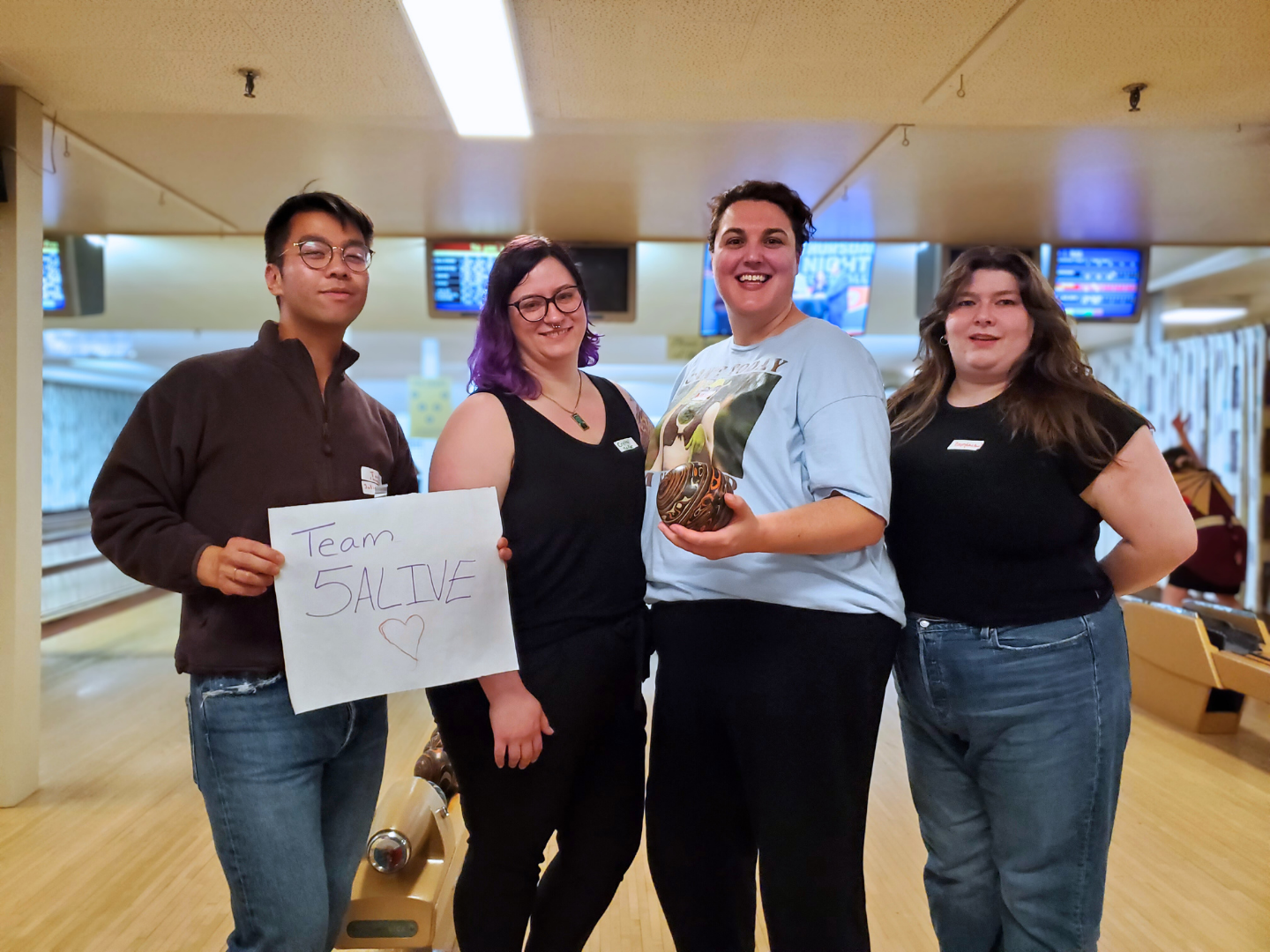 4 bowlers pose for the camera. One is holding a sign that says "Team 5Alive", while another proudly holds a 5 pin bowling ball.