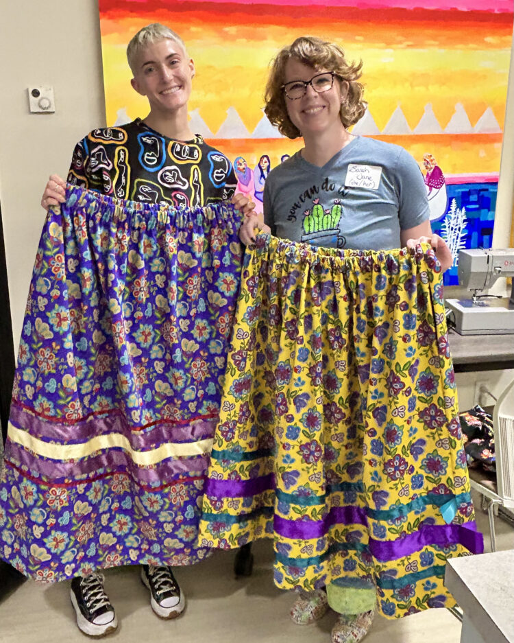 Two smiling blonde facilitators stand proudly showing off their newly-sewn ribbon skirts
